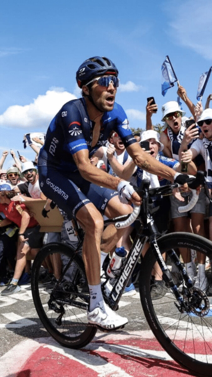 Un cycliste en plein effort est applaudi par une foule enthousiaste, brandissant des drapeaux et prenant des photos.