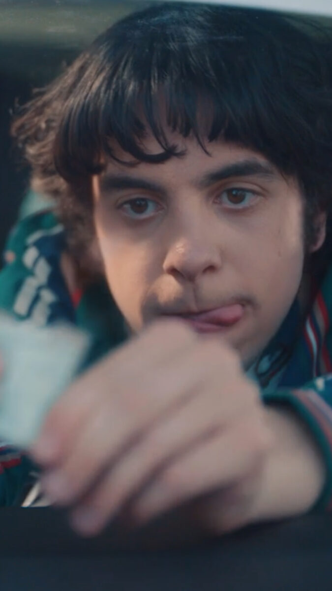 Un jeune homme aux cheveux bouclés apparaît dans une voiture, tirant la langue tout en regardant intensément quelque chose dans sa main.