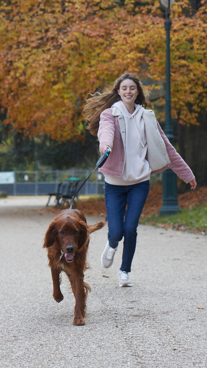 Une jeune femme souriante court en tenant en laisse un chien brun dans un parc aux feuilles d'automne.