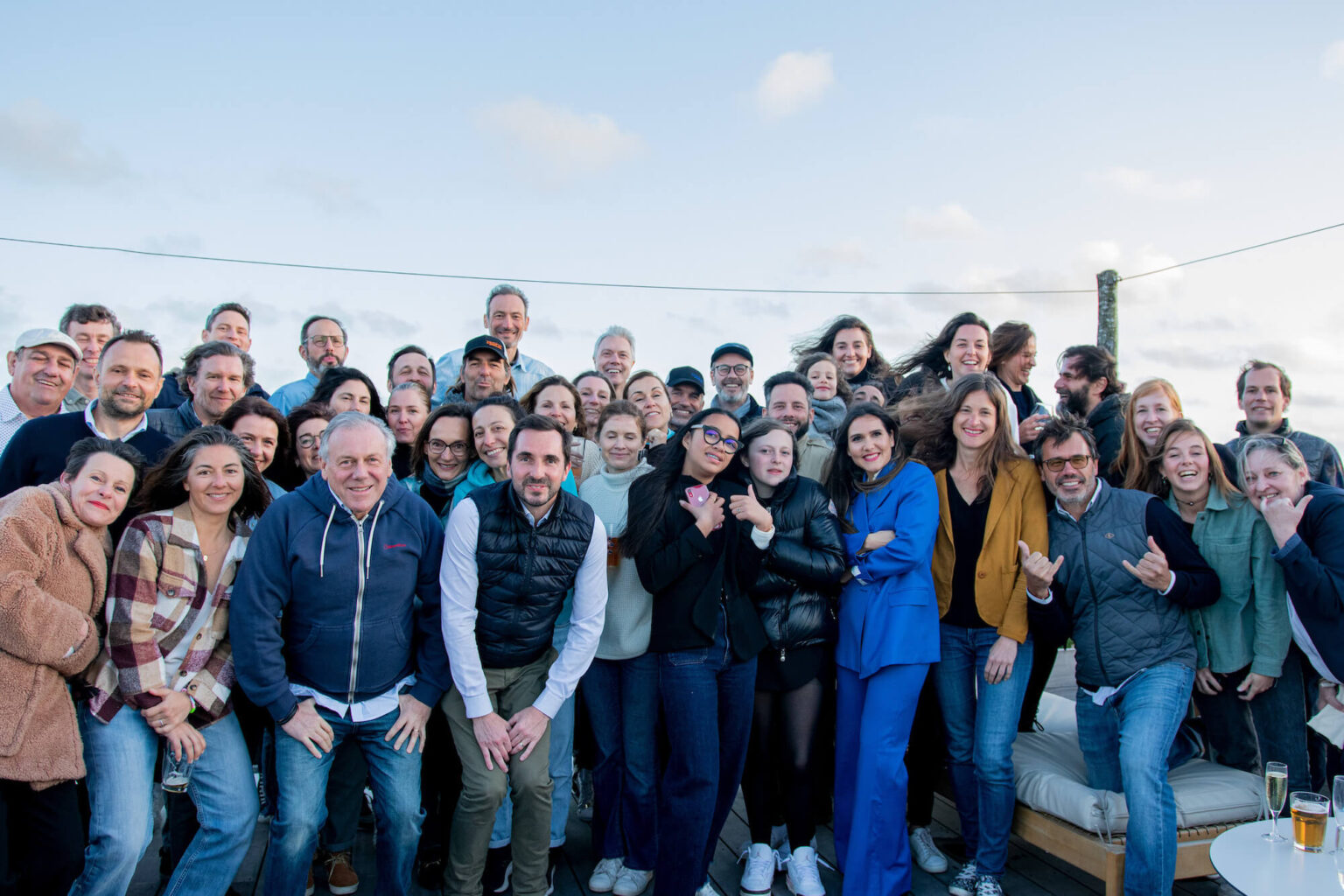 Un grand groupe de personnes souriantes pose ensemble à l'extérieur sous un ciel bleu, créant une atmosphère conviviale.
