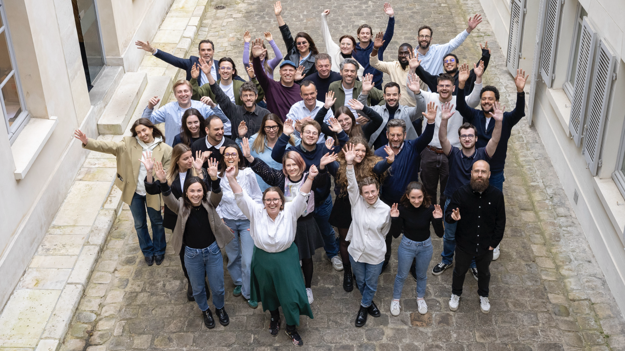 Un groupe de personnes souriantes se tient ensemble dans une ruelle, levant les bras en l'air pour une photo de groupe joyeuse.