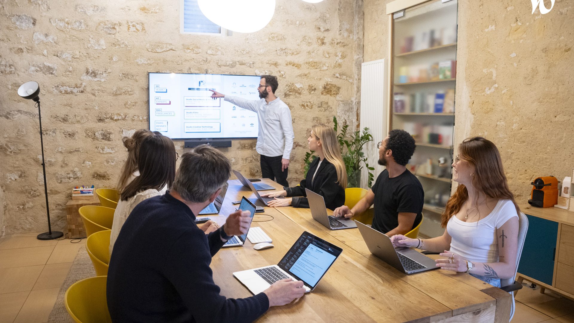 Une réunion d'équipe a lieu autour d'une table en bois, avec plusieurs participants attentifs regardant un écran où un homme présente des informations.