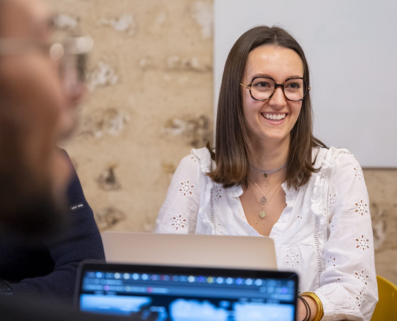 Une femme souriante portant une chemise blanche est assise à une table dans un environnement de bureau, avec un ordinateur portable visible au premier plan.