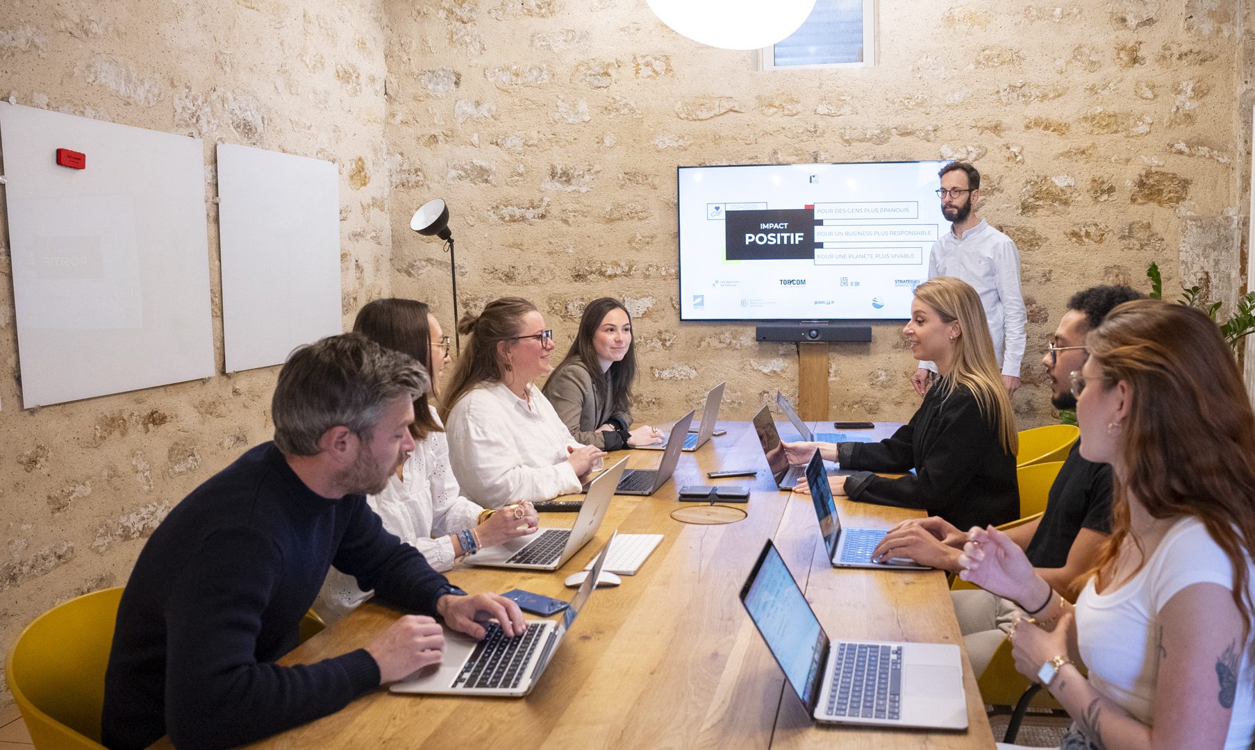 Une équipe de jeunes professionnels discute autour d'une table en bois, avec des ordinateurs portables ouverts, dans une salle de réunion moderne avec des murs en pierre.