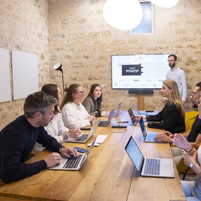 Une équipe de jeunes professionnels discute autour d'une table en bois, avec des ordinateurs portables ouverts, dans une salle de réunion moderne avec des murs en pierre.