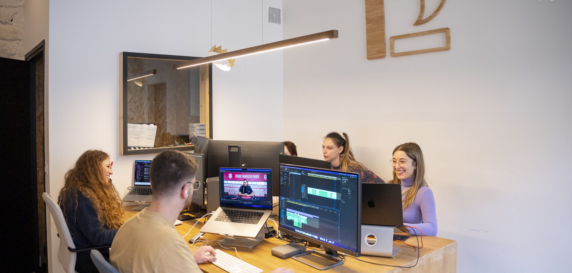 Un groupe de jeunes professionnels travaille dans un bureau moderne et lumineux, avec des ordinateurs et des écrans affichant des contenus numériques.
