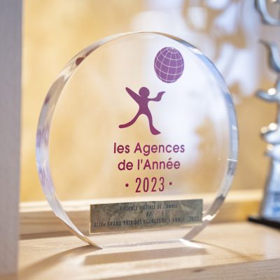 Le visuel montre un prix en verre transparent gravé avec le texte "les Agences de l'Année 2023" et une silhouette en action, accompagné d'autres trophées en arrière-plan.