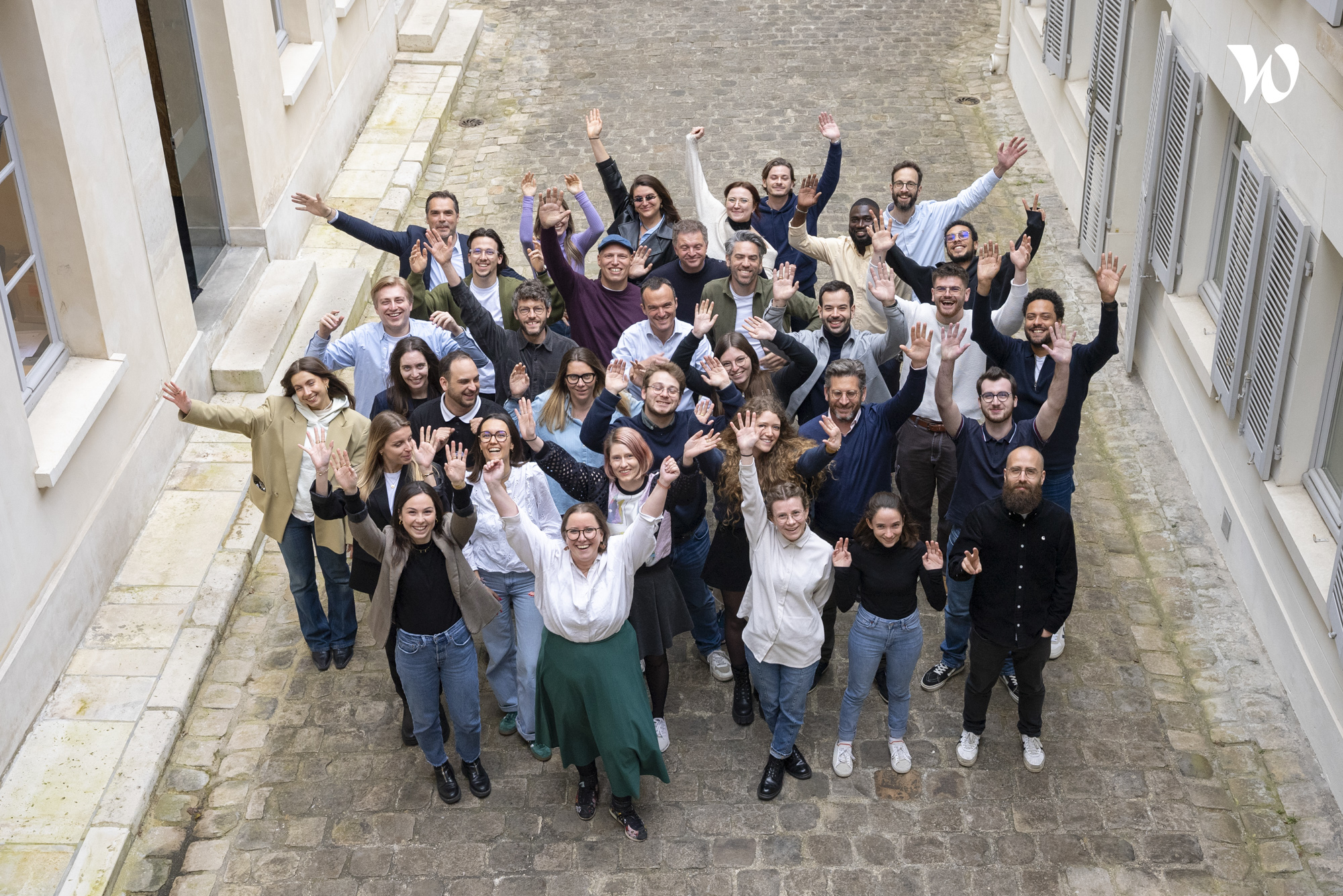 Un groupe de personnes souriantes se tient dans une ruelle pavée, levant les bras en signe de joie.