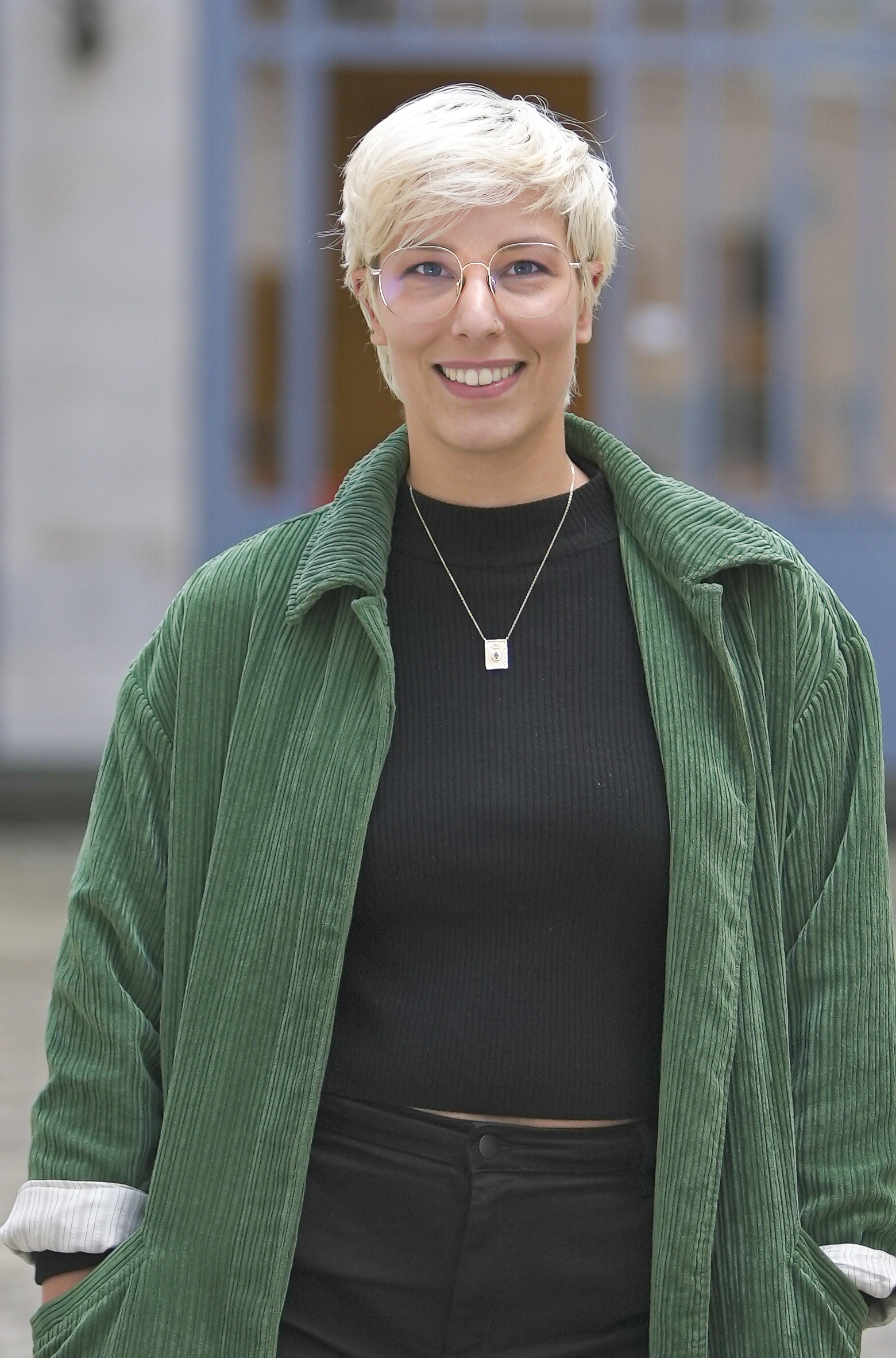 Une personne souriante aux cheveux courts et blonds portant une veste verte et un pull noir se tient devant un fond flou.