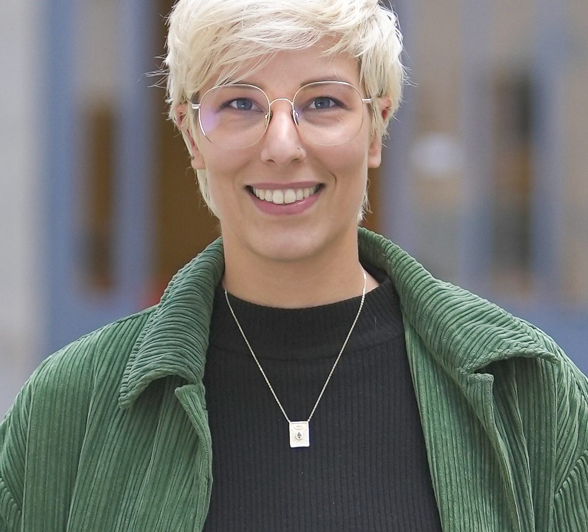 Une personne souriante avec des lunettes et des cheveux courts, portant un manteau vert et un haut noir, se tient devant un bâtiment flou.
