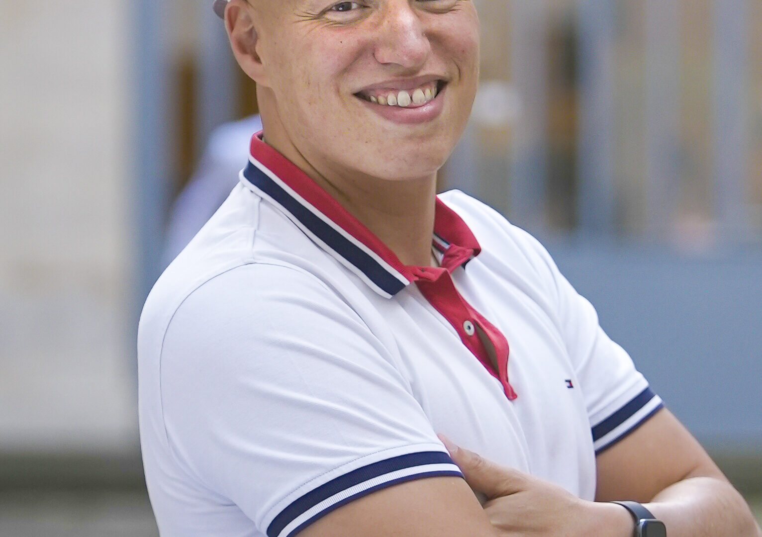 Un homme souriant, portant un polo blanc et une casquette, se tient avec les bras croisés dans une rue.